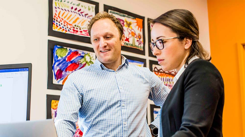 Two colleagues in discussion. They are looking at a laptop, with one smiling and the other colleague looking focused. Behind them is a display of colourful children’s drawings.