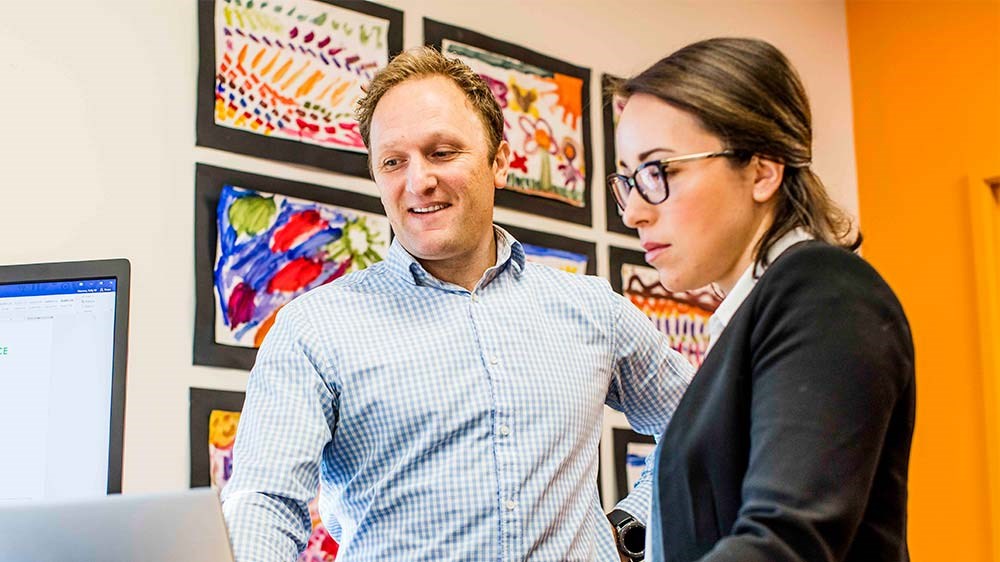 Two colleagues in discussion. They are looking at a laptop, with one smiling and the other colleague looking focused. Behind them is a display of colourful children’s drawings.