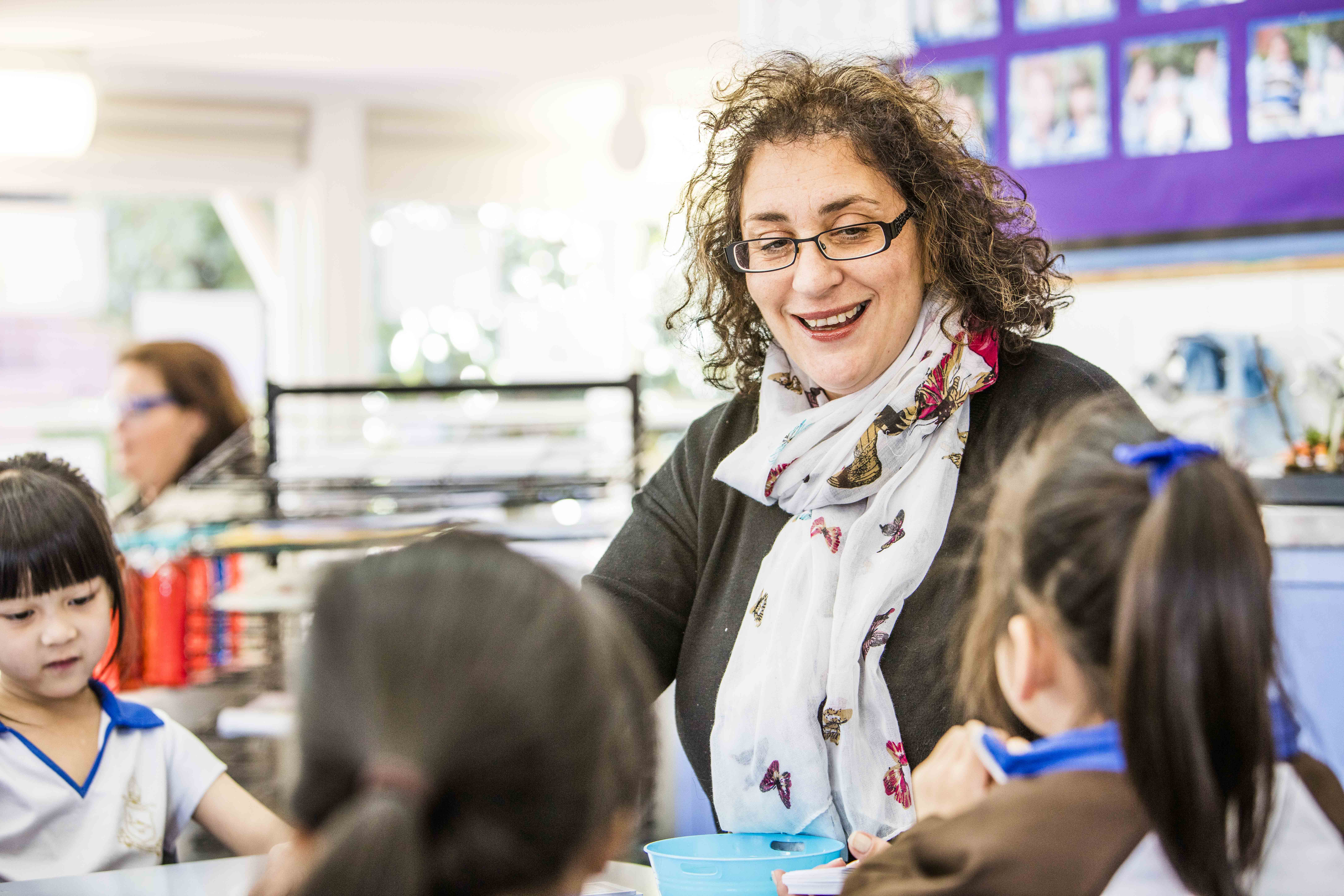 Teacher sitting with children at a large, low desk. The teacher has a small blue bucket in front of her. Behind the teacher is an art drying rack, books, and a noticeboard decorated with photographs. 