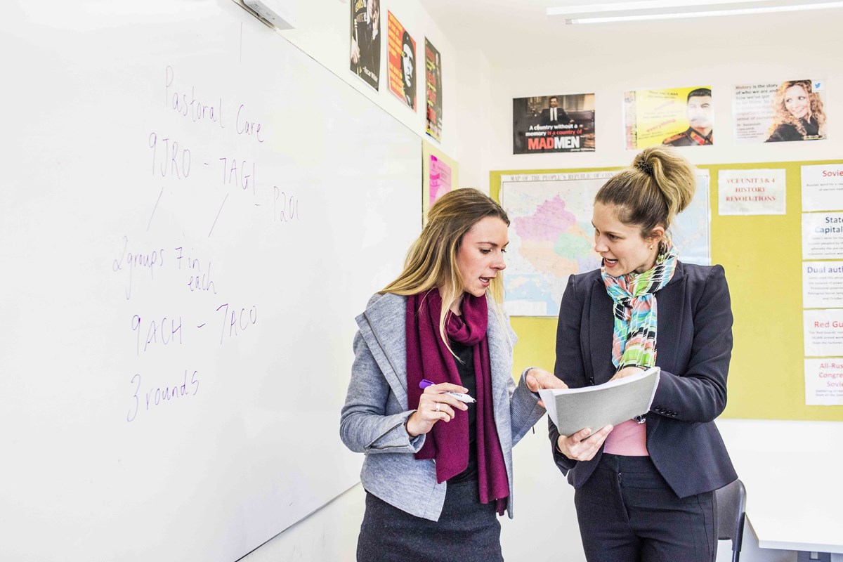 Two teachers discuss in front of a whiteboard. The teacher on the left is holding a whiteboard marker. The other teacher is holding a stack of papers that both of them are referring to.
