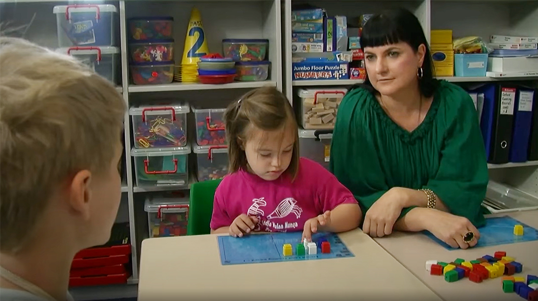 A primary school teacher in a pre-primary/prep and year 1 room sits with two learners. One learner is using blocks in a learning task, whilst the other is sitting across from the teacher.