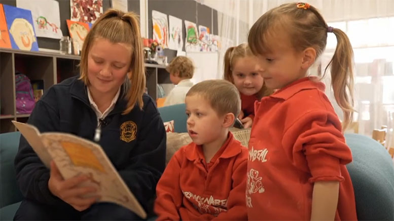 Three young learners are gathered around a secondary school student who is reading with them.