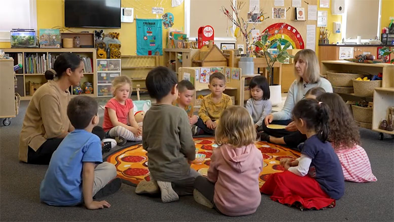Two teachers sit on the floor in a circle with 10 young learners in a pre-primary/preschool learning environment. They are engaging in an activity using toy pizzas.