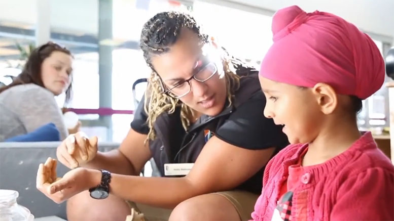 A teacher works with a young learner in the foreground, with another educator watching them in the background.
